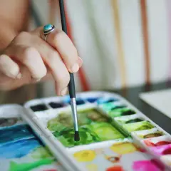 Main d'une artiste avec une bague turquoise, tenant un pinceau au-dessus d'une palette de couleurs vives pour une aquarelle peinture.