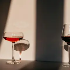Deux verres de vin, l'un rouge clair, l'autre plus foncé, se dressent dans la lumière. Leurs ombres dansent sur le mur, évoquant l'origine de l'eau.
