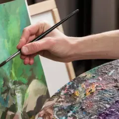 Main tenant une palette colorée, une main fine utilise un pinceau pour peindre à l'acrylique sur une toile verte.