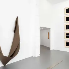 Une installation d'art minimaliste dans une galerie blanche. Une sculpture textile drapée à gauche, des œuvres encadrées à droite.