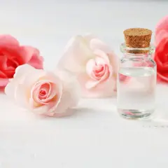Un flacon de verre bouché de liège contient un liquide clair, entouré de roses roses et pâles. L'expression de l'eau de rose évoque la douceur et la pureté.