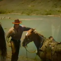 Un cow-boy, prêt à monter sur ses grands chevaux, caresse son compagnon près d'une rivière.