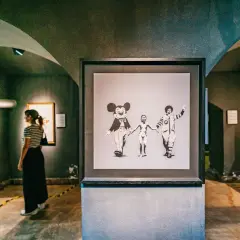 Dans une galerie, une femme observe une œuvre de street art connu, Banksy, représentant Mickey, un enfant nu et Ronald McDonald se tenant la main.