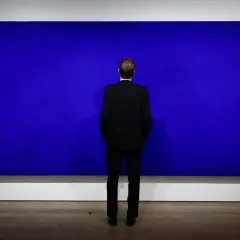 Un homme en costume noir contemple une immense peinture monochrome bleu, une œuvre qui invite à la méditation.