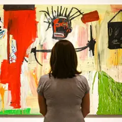 Une femme admire une peinture vibrante dans un style Basquiat, avec un visage expressif au centre et des formes abstraites.