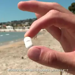 Main d'un homme tenant une pilule blanche sur une plage ensoleillée, interrogeant sur l'expression "se dorer la pilule".