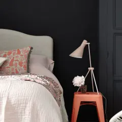 Chambre sombre avec lit douillet, coussin à motifs, lampe rose et un tabouret orange rappelant une camera obscura.
