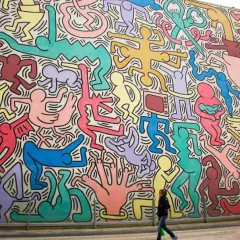 Murale coloré de figures humaines stylisées, typiques des œuvres d'art de Keith Haring, envahissant un bâtiment. Une femme passe devant.