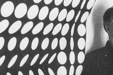 Un homme pose devant une œuvre op art de Vasarely, un motif de cercles blancs sur fond noir créant une illusion de mouvement.