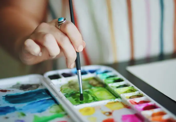Main d'une artiste avec une bague turquoise, tenant un pinceau au-dessus d'une palette de couleurs vives pour une aquarelle peinture.