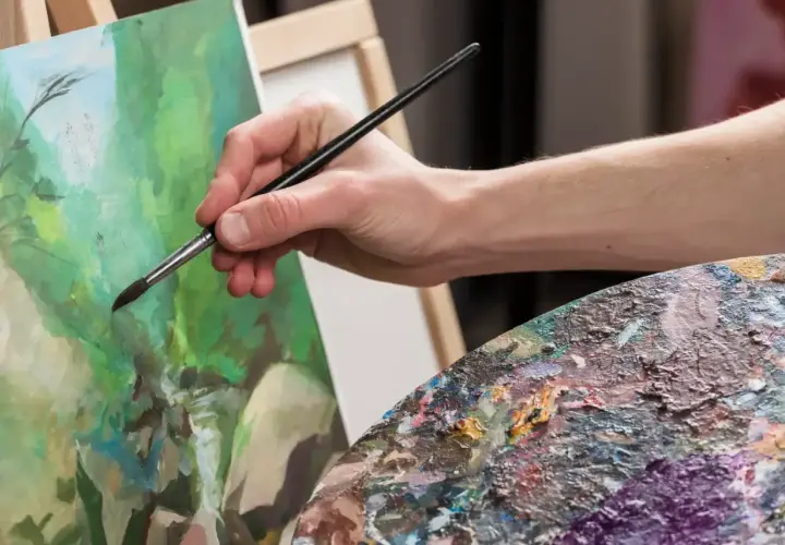 Main tenant une palette colorée, une main fine utilise un pinceau pour peindre à l'acrylique sur une toile verte.