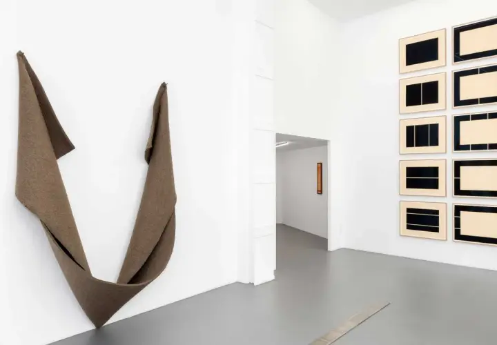 Une installation d'art minimaliste dans une galerie blanche. Une sculpture textile drapée à gauche, des œuvres encadrées à droite.