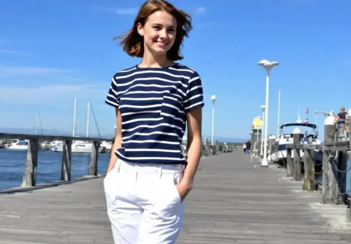 Jeune femme souriante sur un ponton, portant un t-shirt rayé marine et un pantalon blanc. L'ambiance est estivale et décontractée.