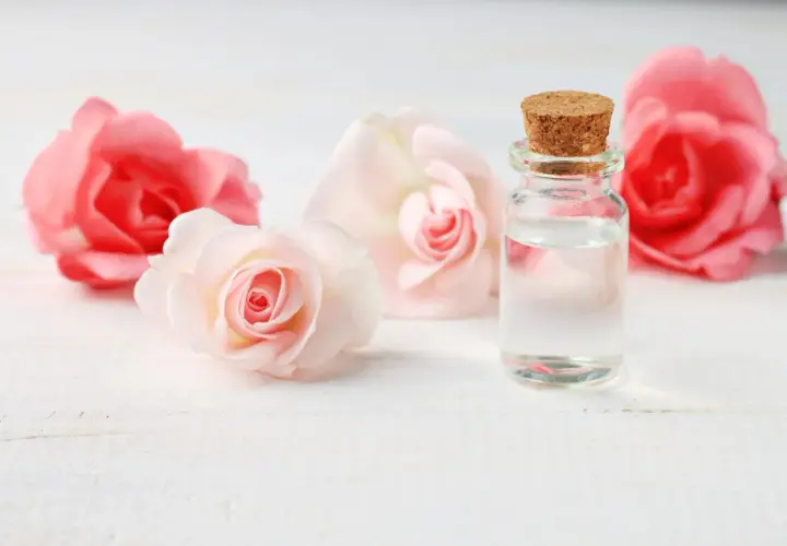 Un flacon de verre bouché de liège contient un liquide clair, entouré de roses roses et pâles. L'expression de l'eau de rose évoque la douceur et la pureté.