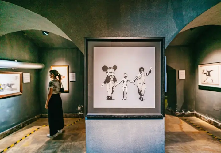 Dans une galerie, une femme observe une œuvre de street art connu, Banksy, représentant Mickey, un enfant nu et Ronald McDonald se tenant la main.