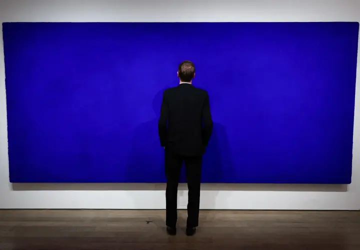 Un homme en costume noir contemple une immense peinture monochrome bleu, une œuvre qui invite à la méditation.