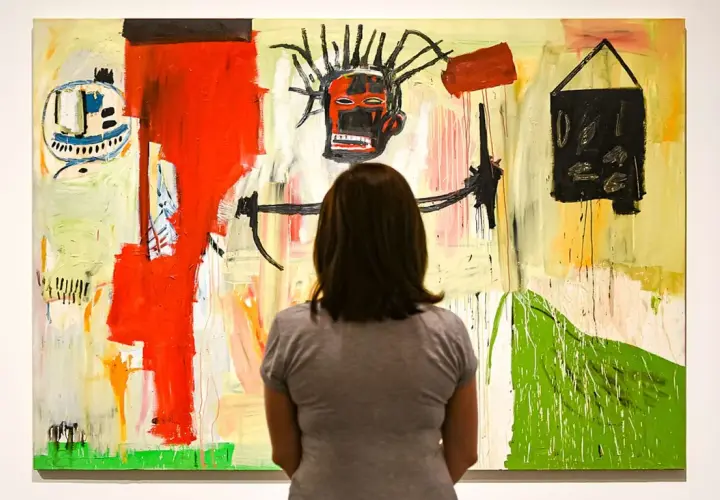 Une femme admire une peinture vibrante dans un style Basquiat, avec un visage expressif au centre et des formes abstraites.
