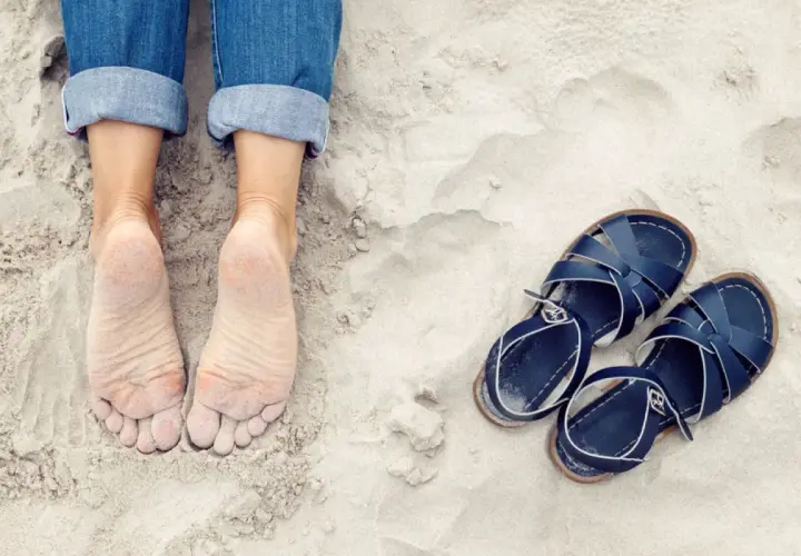 Pieds nus sur le sable, jeans retroussés, et une paire de sandales bleues à côté. On dirait que quelqu'un est prêt à être à côté de ses pompes pour une promenade.