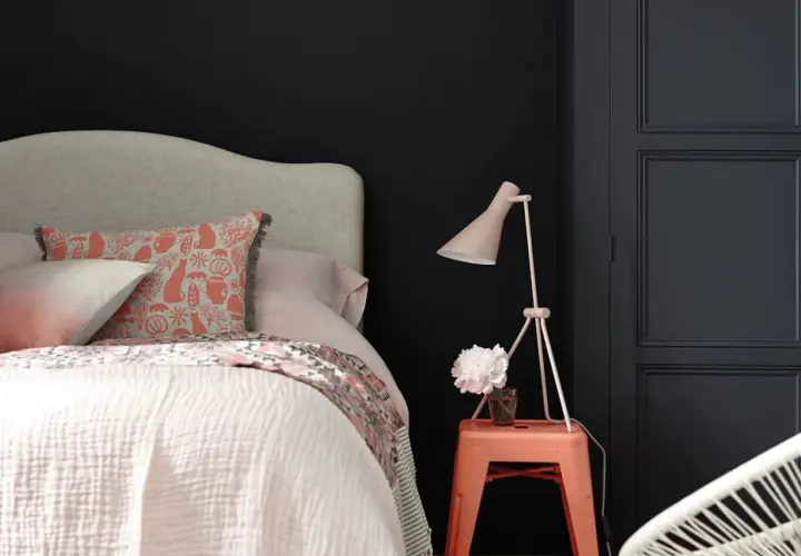 Chambre sombre avec lit douillet, coussin à motifs, lampe rose et un tabouret orange rappelant une camera obscura.