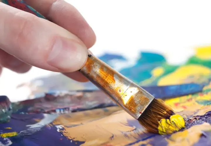 Main d'artiste saisissant un pinceau chargé de peinture jaune, prêt à créer un tableau peinture à l'huile facile sur une palette colorée.