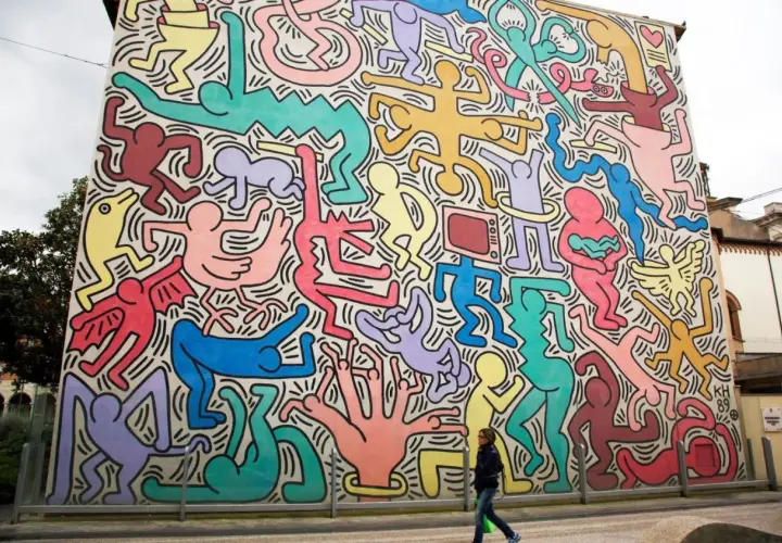 Murale coloré de figures humaines stylisées, typiques des œuvres d'art de Keith Haring, envahissant un bâtiment. Une femme passe devant.