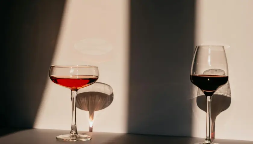 Deux verres de vin, l'un rouge clair, l'autre plus foncé, se dressent dans la lumière. Leurs ombres dansent sur le mur, évoquant l'origine de l'eau.