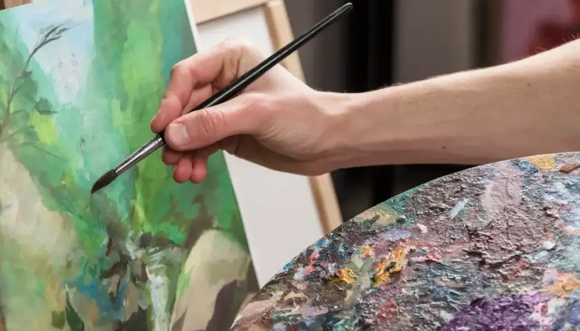 Main tenant une palette colorée, une main fine utilise un pinceau pour peindre à l'acrylique sur une toile verte.