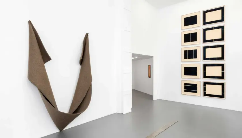 Une installation d'art minimaliste dans une galerie blanche. Une sculpture textile drapée à gauche, des œuvres encadrées à droite.