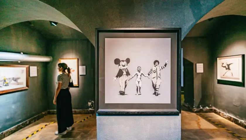Dans une galerie, une femme observe une œuvre de street art connu, Banksy, représentant Mickey, un enfant nu et Ronald McDonald se tenant la main.