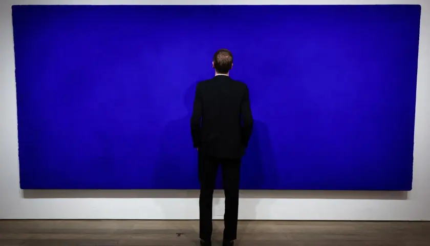 Un homme en costume noir contemple une immense peinture monochrome bleu, une œuvre qui invite à la méditation.