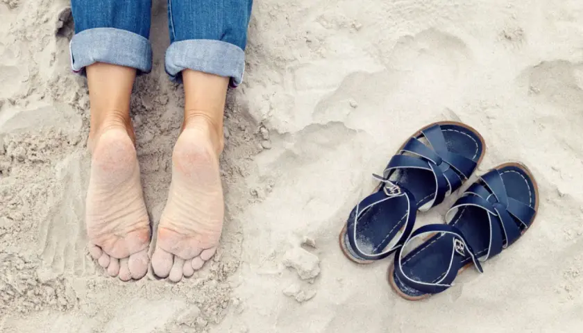 Pieds nus sur le sable, jeans retroussés, et une paire de sandales bleues à côté. On dirait que quelqu'un est prêt à être à côté de ses pompes pour une promenade.