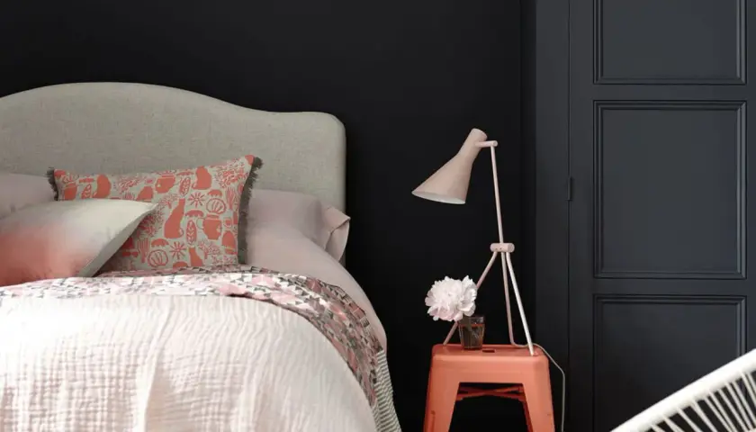 Chambre sombre avec lit douillet, coussin à motifs, lampe rose et un tabouret orange rappelant une camera obscura.
