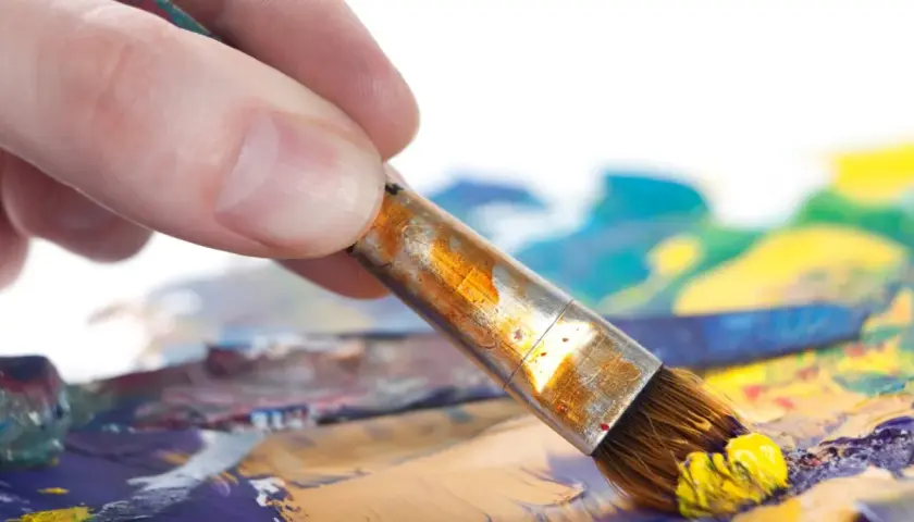 Main d'artiste saisissant un pinceau chargé de peinture jaune, prêt à créer un tableau peinture à l'huile facile sur une palette colorée.