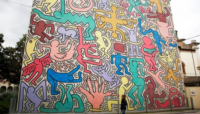 Murale coloré de figures humaines stylisées, typiques des œuvres d'art de Keith Haring, envahissant un bâtiment. Une femme passe devant.