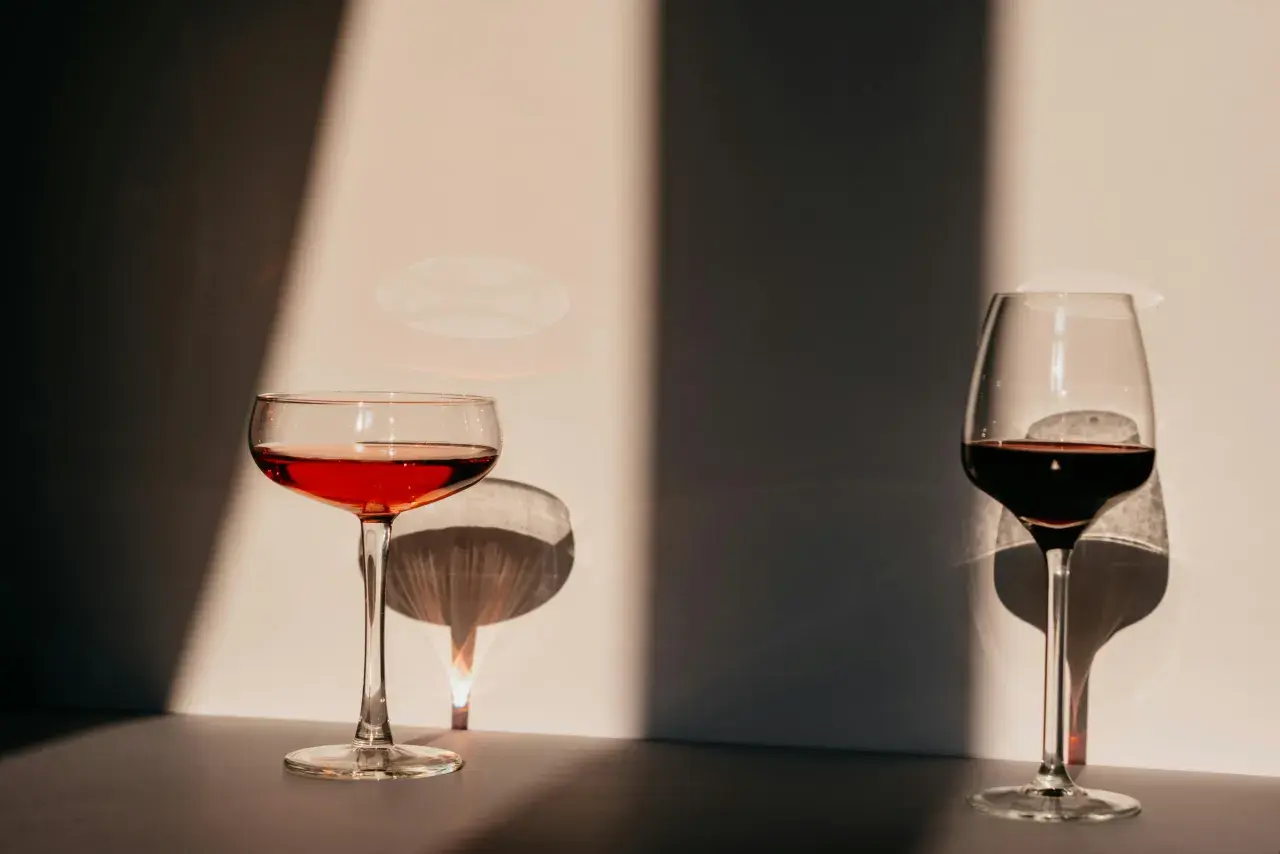 Deux verres de vin, l'un rouge clair, l'autre plus foncé, se dressent dans la lumière. Leurs ombres dansent sur le mur, évoquant l'origine de l'eau.
