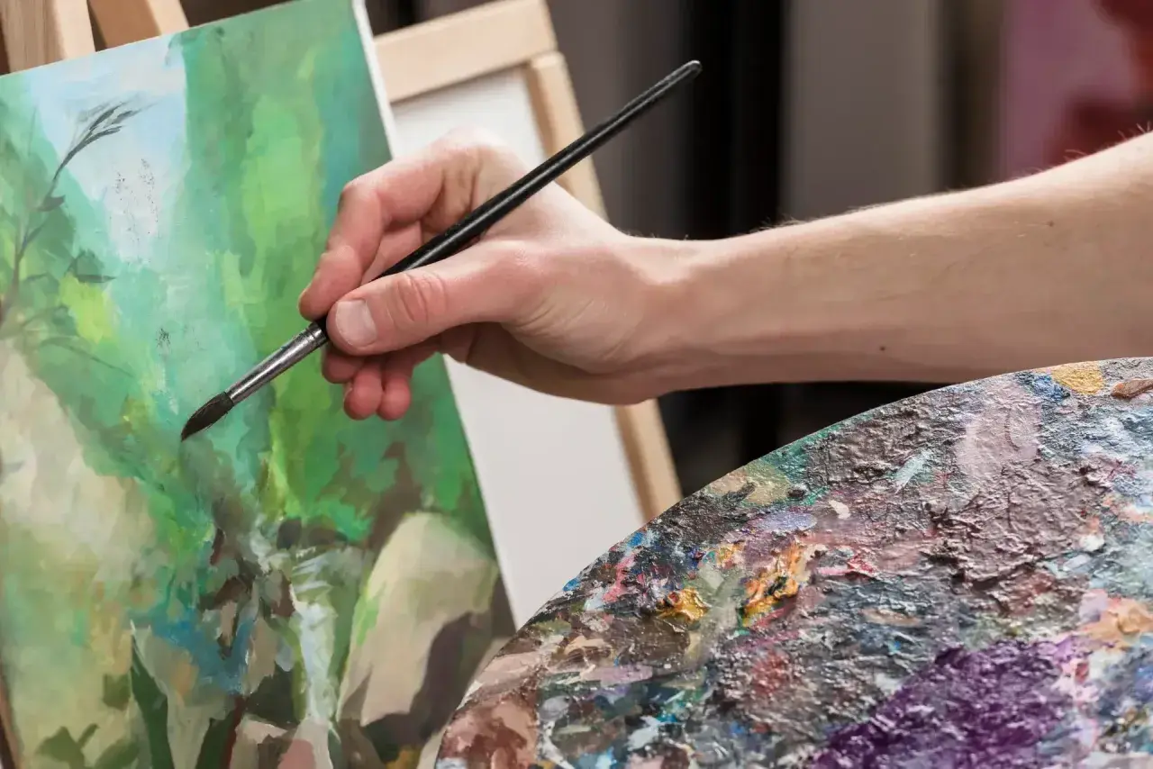 Main tenant une palette colorée, une main fine utilise un pinceau pour peindre à l'acrylique sur une toile verte.