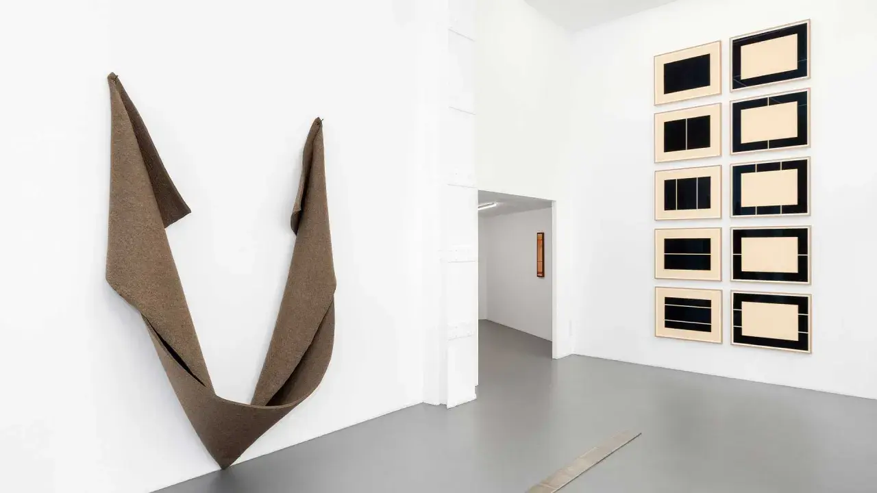 Une installation d'art minimaliste dans une galerie blanche. Une sculpture textile drapée à gauche, des œuvres encadrées à droite.
