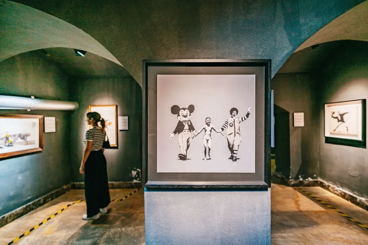 Dans une galerie, une femme observe une œuvre de street art connu, Banksy, représentant Mickey, un enfant nu et Ronald McDonald se tenant la main.