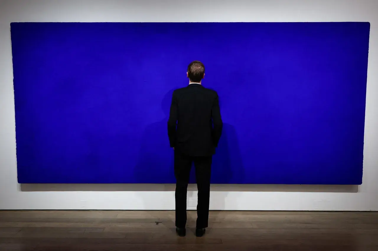 Un homme en costume noir contemple une immense peinture monochrome bleu, une œuvre qui invite à la méditation.