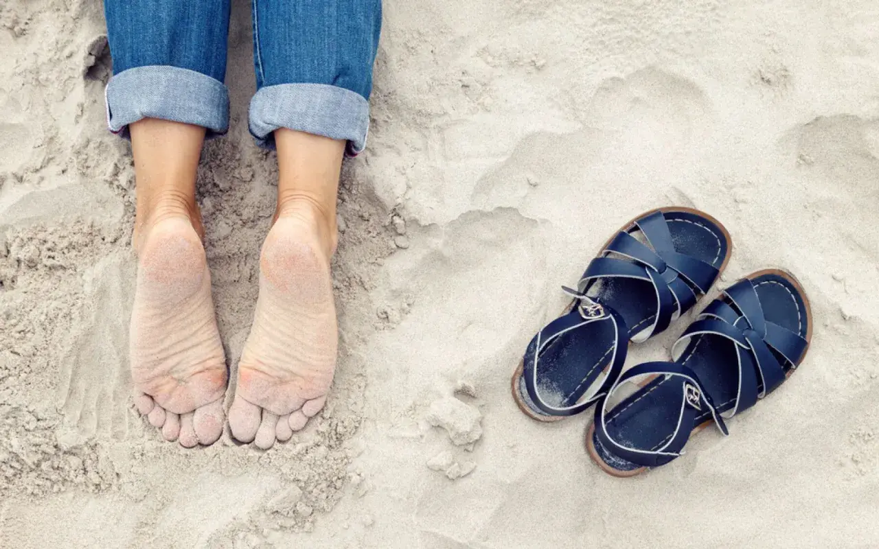 Pieds nus sur le sable, jeans retroussés, et une paire de sandales bleues à côté. On dirait que quelqu'un est prêt à être à côté de ses pompes pour une promenade.