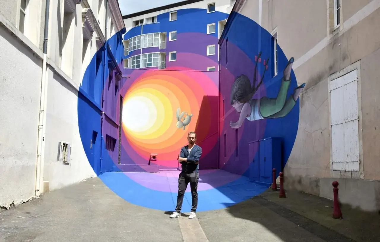 Un artiste street art pose devant son œuvre : un tunnel coloré avec une fillette et un lapin flottant.