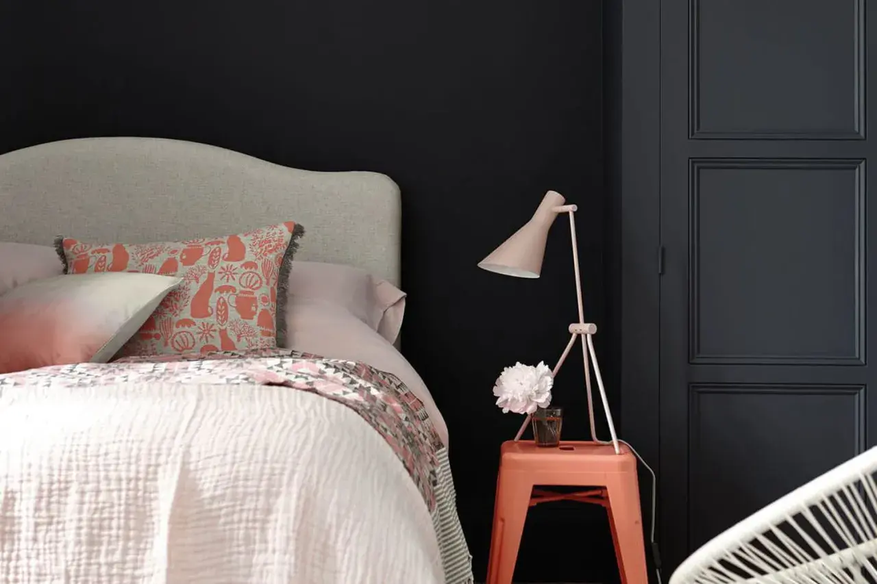 Chambre sombre avec lit douillet, coussin à motifs, lampe rose et un tabouret orange rappelant une camera obscura.