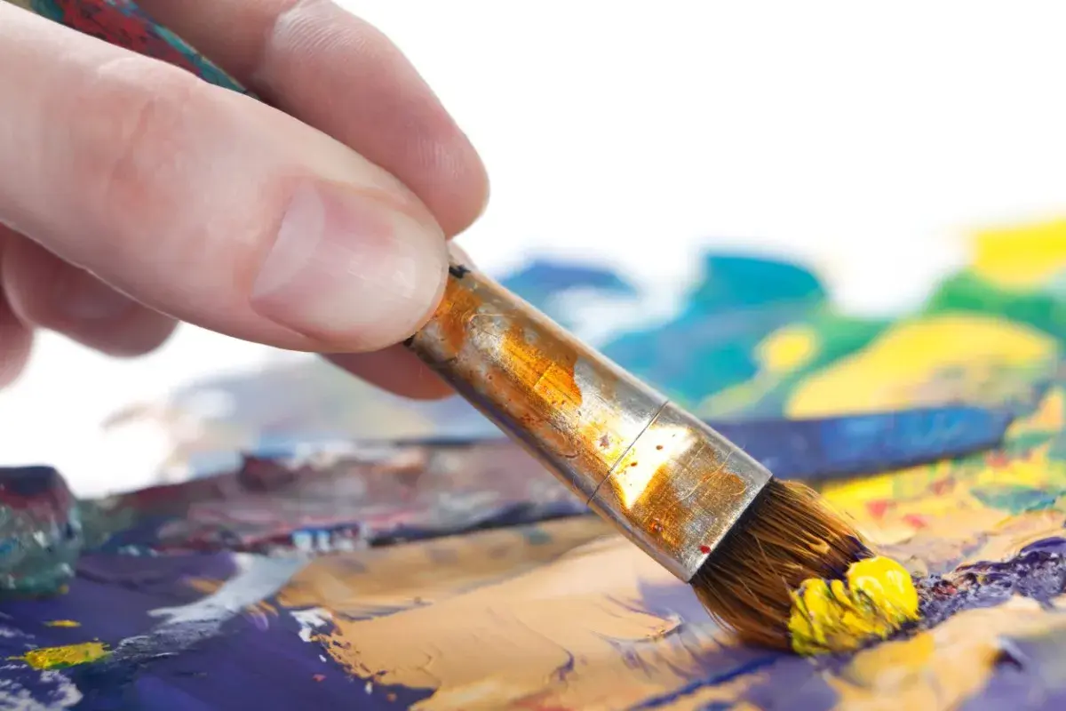 Main d'artiste saisissant un pinceau chargé de peinture jaune, prêt à créer un tableau peinture à l'huile facile sur une palette colorée.