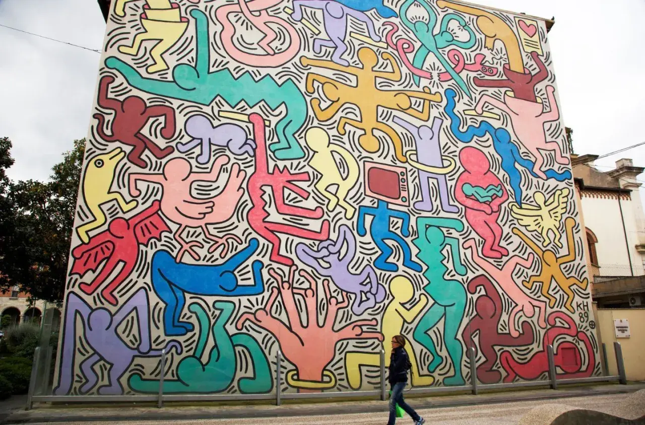 Murale coloré de figures humaines stylisées, typiques des œuvres d'art de Keith Haring, envahissant un bâtiment. Une femme passe devant.