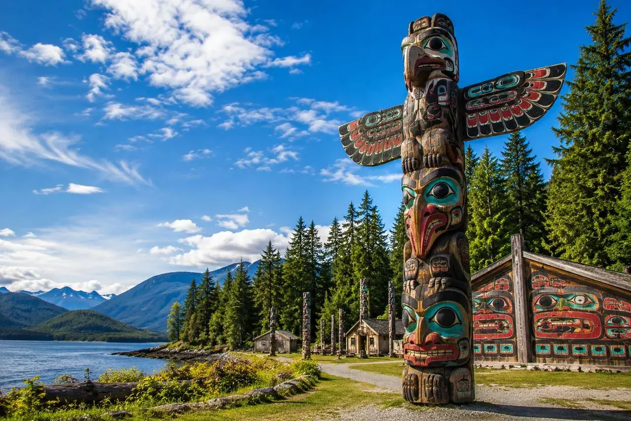Un voyage dans l'art amérindien : un totem coloré et des maisons sculptées se dressent devant un lac et des montagnes majestueuses.