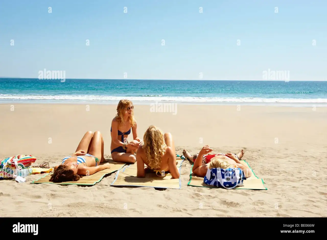 Quatre amies profitent du soleil sur la plage, allongées sur des nattes pour se faire dorer la pilule. L'océan bleu s'étend à l'horizon.