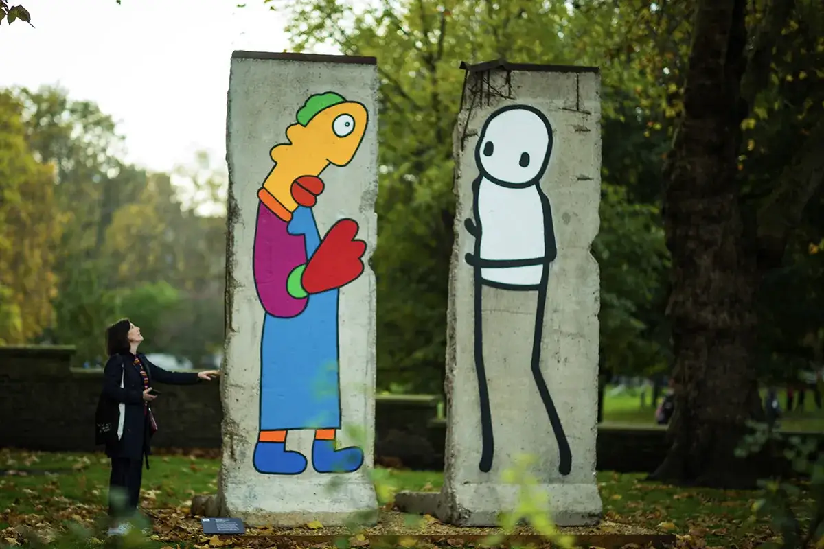 Un fragment du mur de Berlin, peint avec des personnages colorés, face à une femme admirative. L'œuvre évoque Thierry Noir et son art iconique.