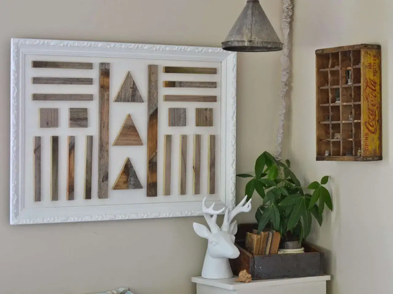 Une oeuvre d'art maison en bois recycl&eacute;, compos&eacute;e de formes g&eacute;om&eacute;triques, encadr&eacute;e de blanc. Une t&ecirc;te de cerf et une plante compl&egrave;tent la sc&egrave;ne.