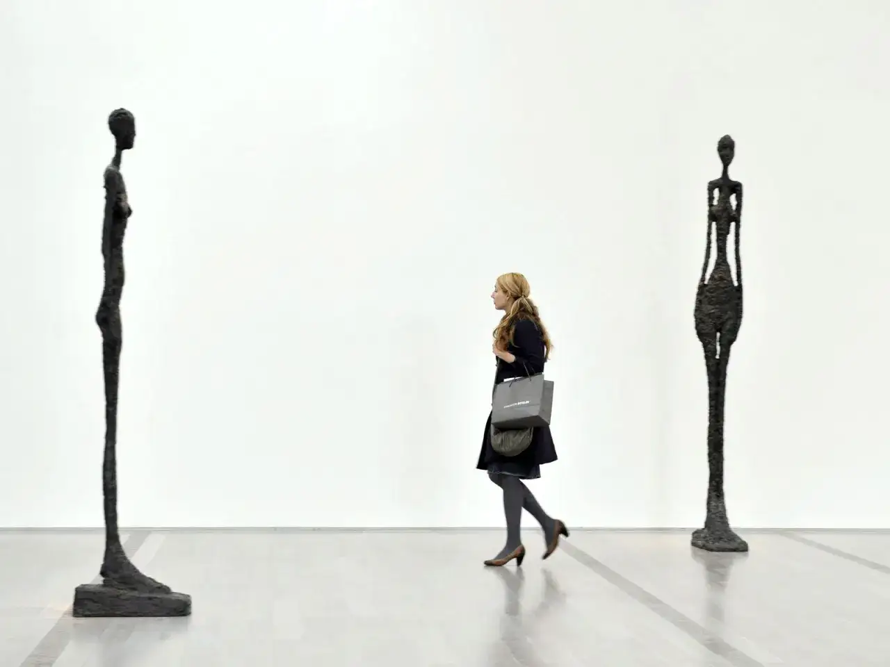 Une femme passe devant deux silhouettes filiformes, des sculptures emblématiques de Giacometti, dans une galerie d'art épurée.